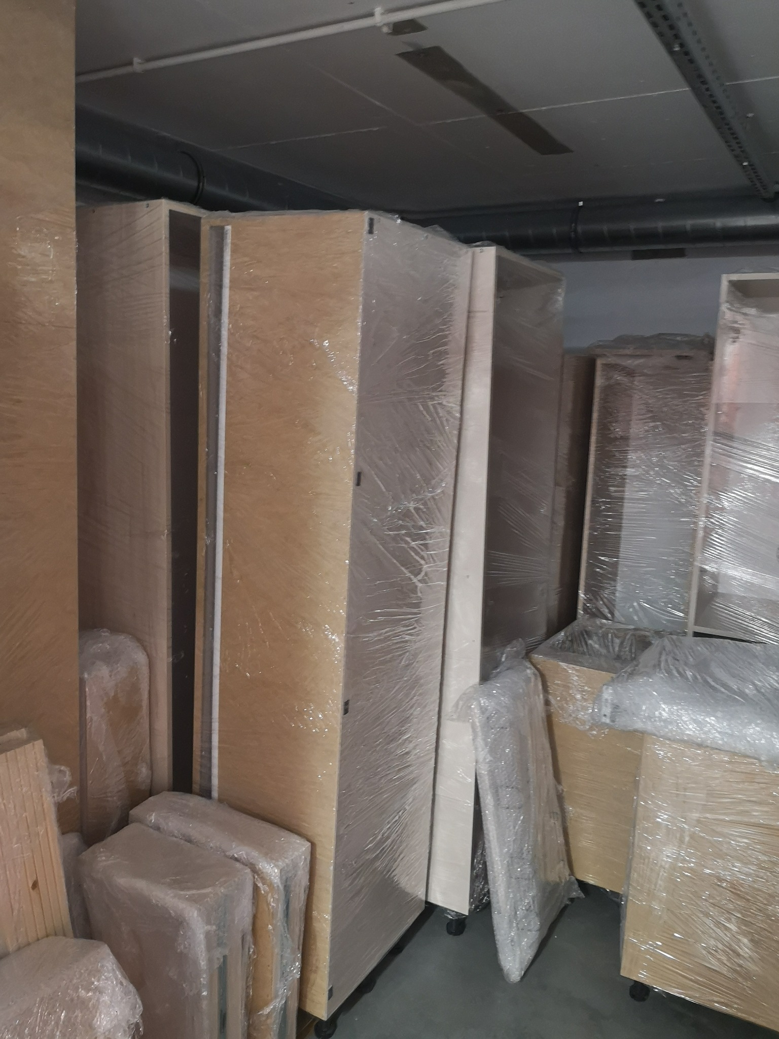 Meblowe elementy zabezpieczone folią stretch, przygotowane do transportu, widoczne kółka transportowe i drewniane elementy, oświetlenie magazynowe.