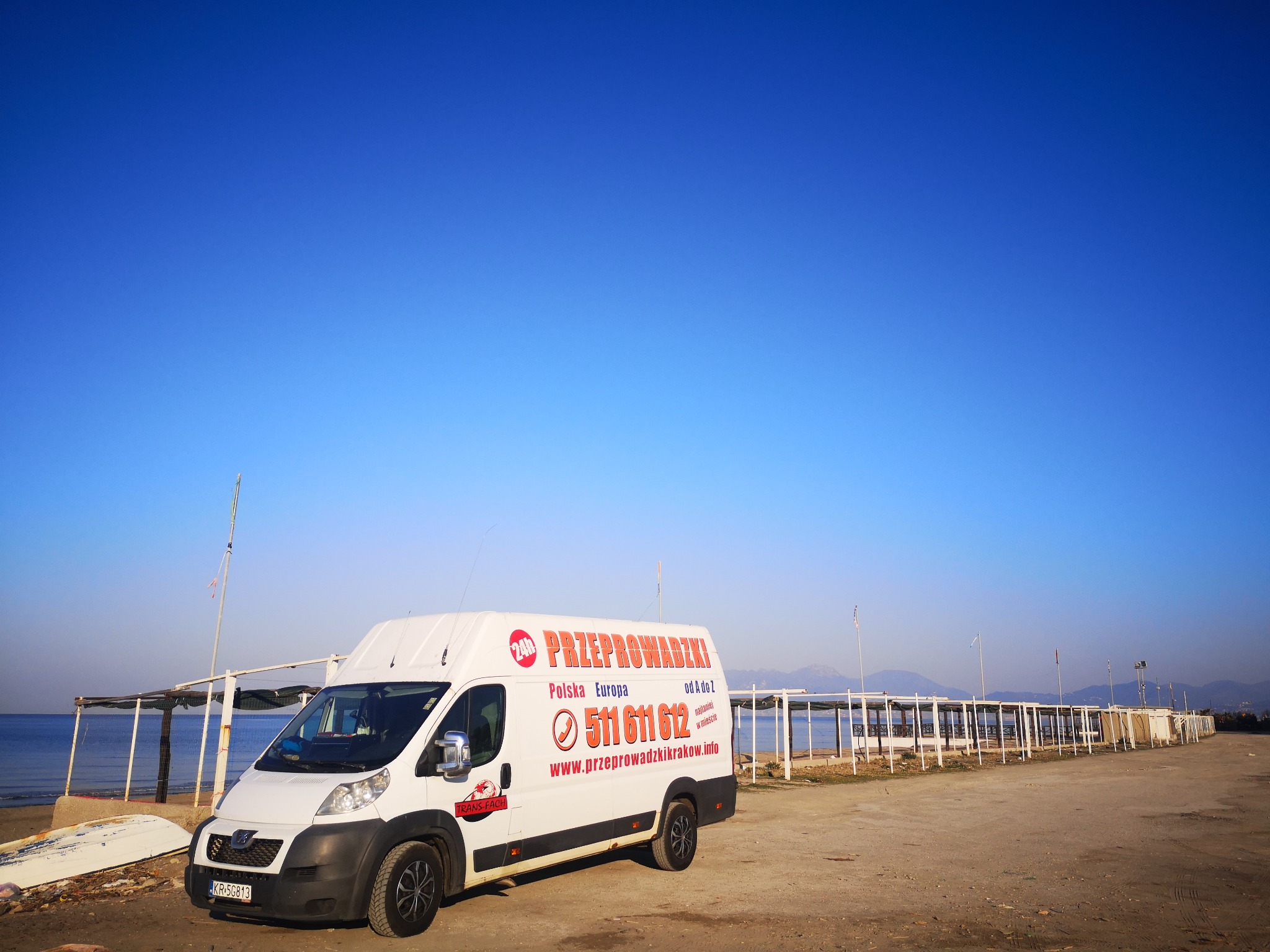 Biały bus z napisem 'Przeprowadzki Polska Europa 511 611 612' zaparkowany na piaszczystej plaży, w tle błękitne niebo i morze z konstrukcjami plażowymi.