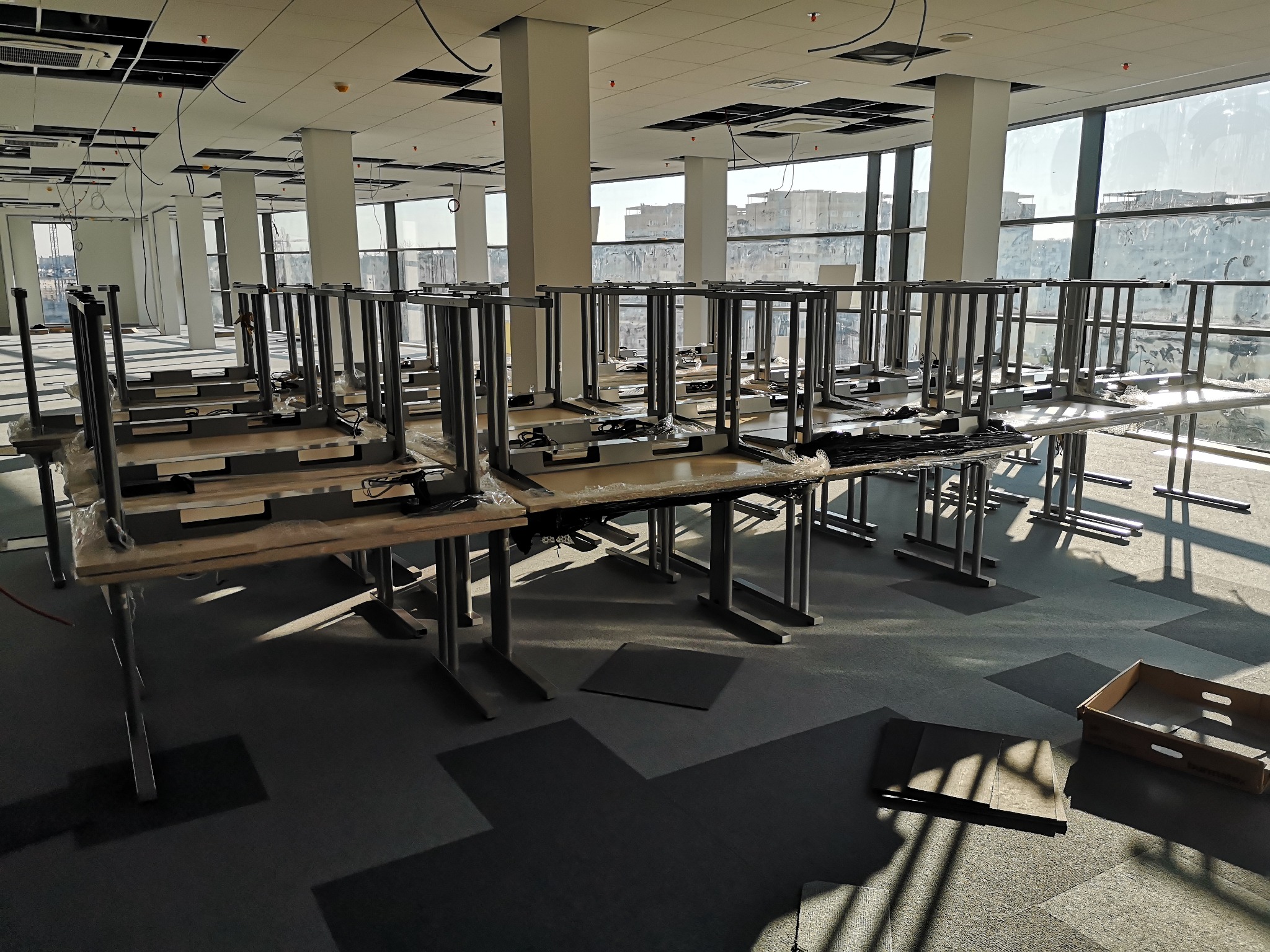 Półprzezroczyste biurko z metalową konstrukcją ustawione do góry nogami w przestronnym biurze z dużymi oknami, widoczne niedokończone prace wykończeniowe sufitu i podłogi.