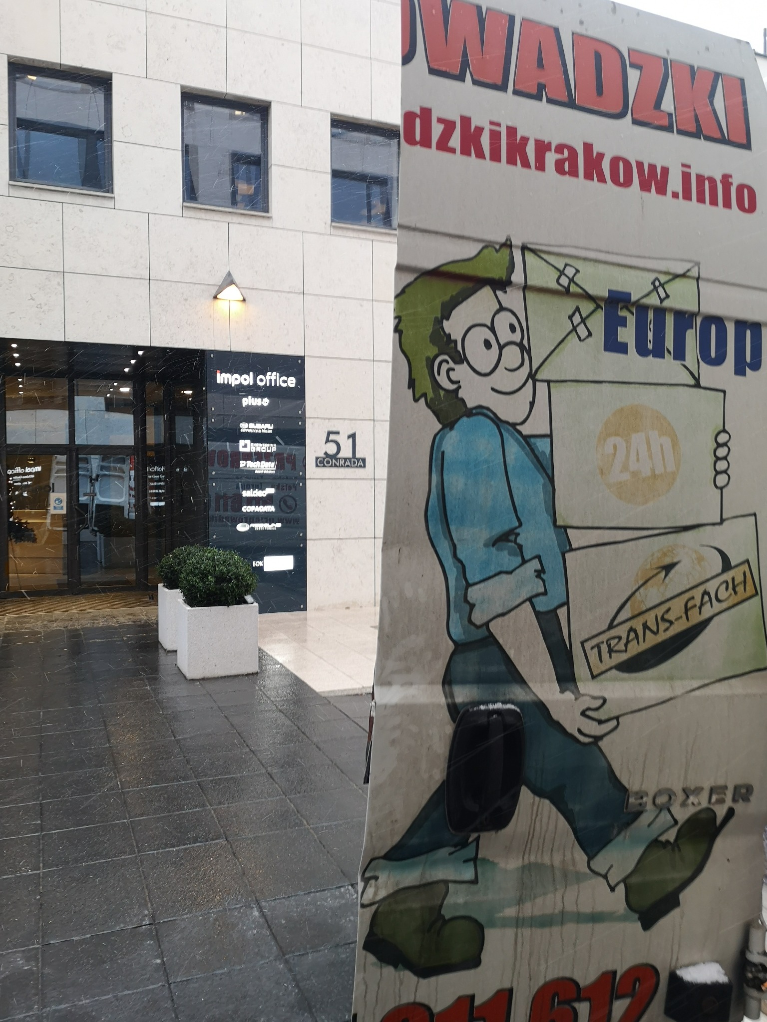 Reklama firmy transportowej przedstawiająca rysunkową postać dźwigającą paczki z napisem 'Europa 24h Trans-Fach Boxer', na tle budynku biurowego Impol Office w Krakowie.
