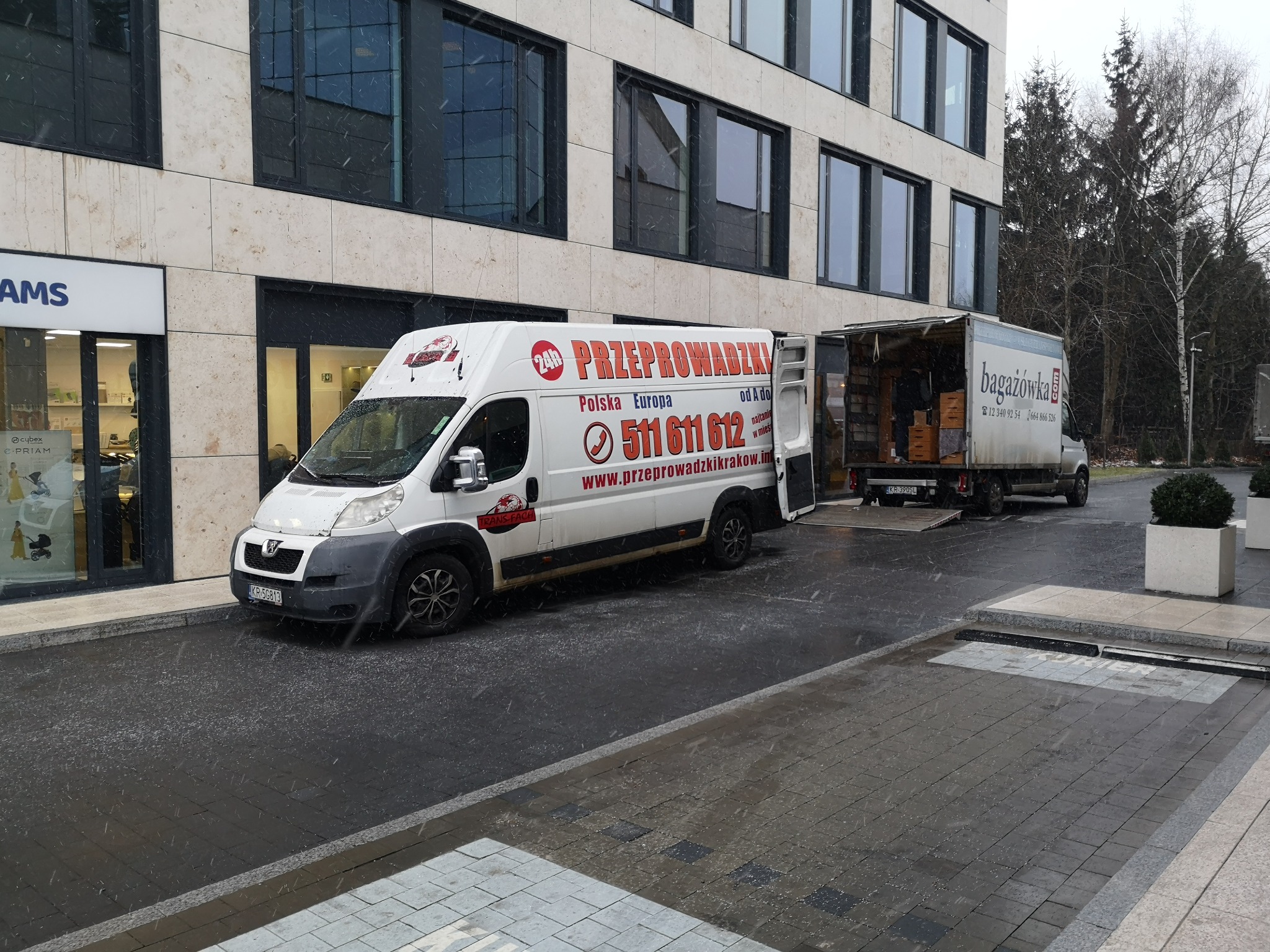 Dwa busy transportowe zaparkowane przed nowoczesnym budynkiem w trakcie opadów śniegu, jeden z otwartą przestrzenią ładunkową wypełnioną kartonami