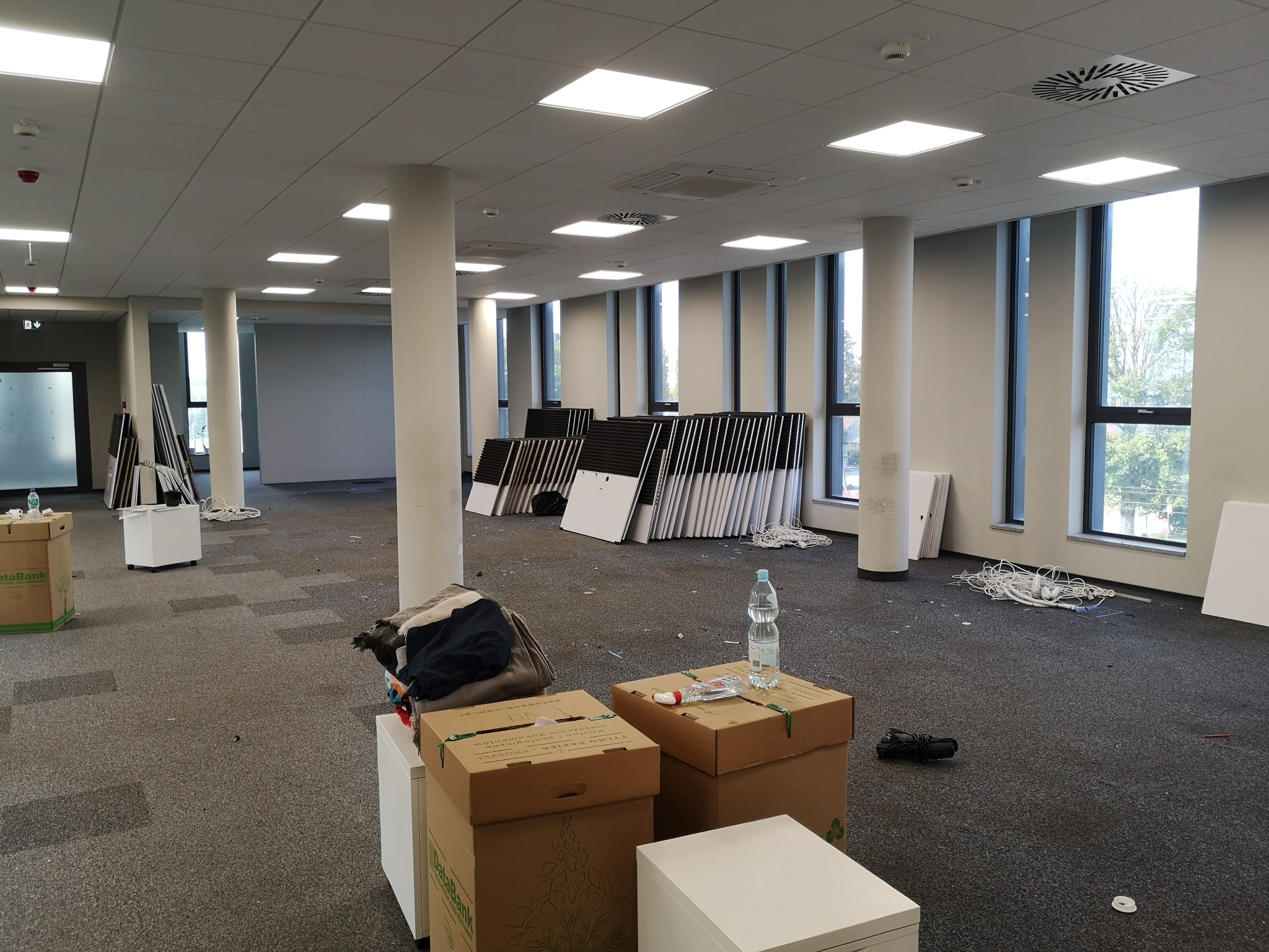 Przestronne biuro po przeprowadzce, z kartonami DataBank, stertą krzeseł biurowych i plątaniną kabli na podłodze z wykładziną w kratkę.
