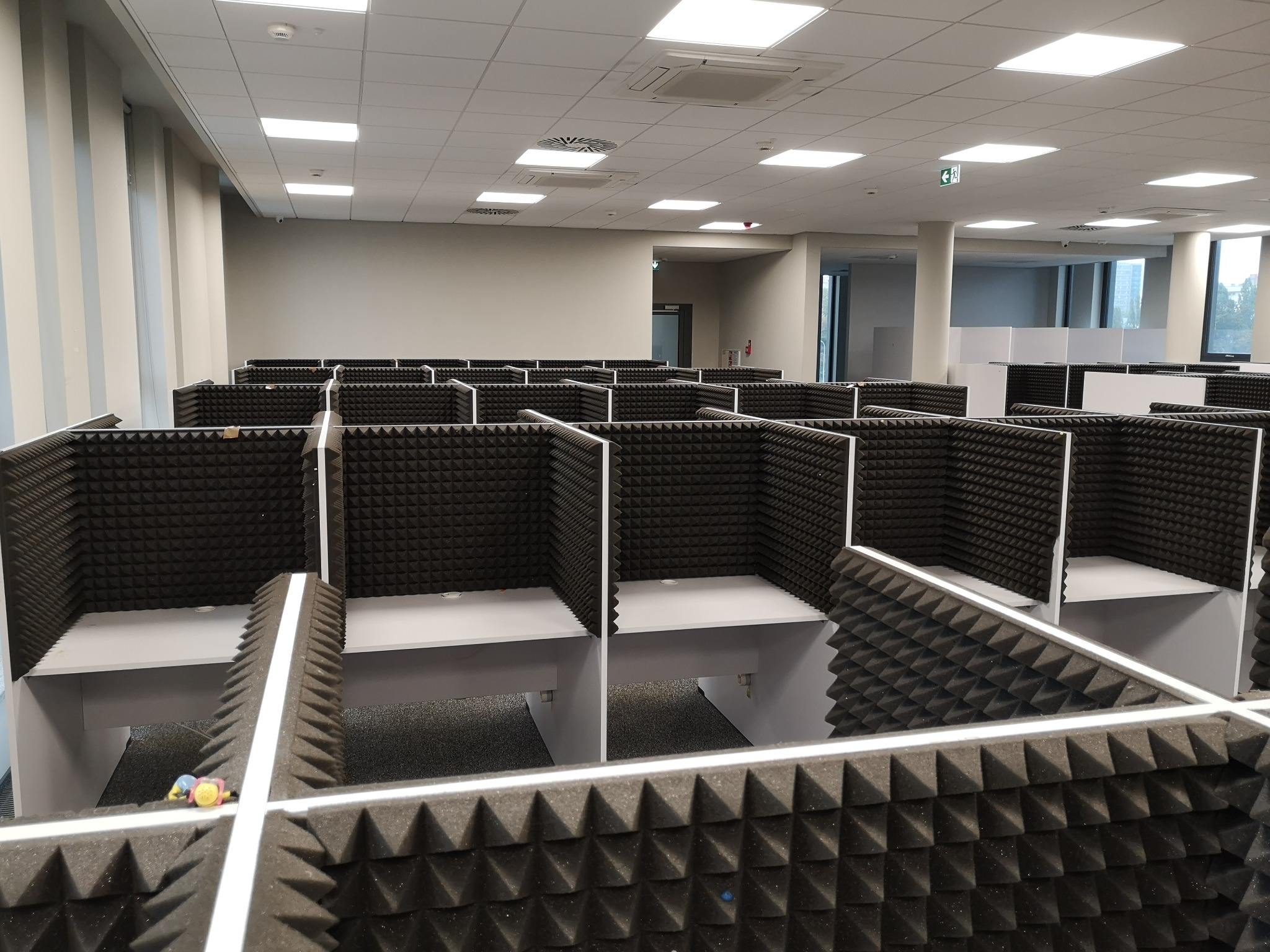 Rząd identycznych, wyciszonych boksów biurowych z szarej pianki akustycznej, widok z góry, w tle jasne biuro z oknami.