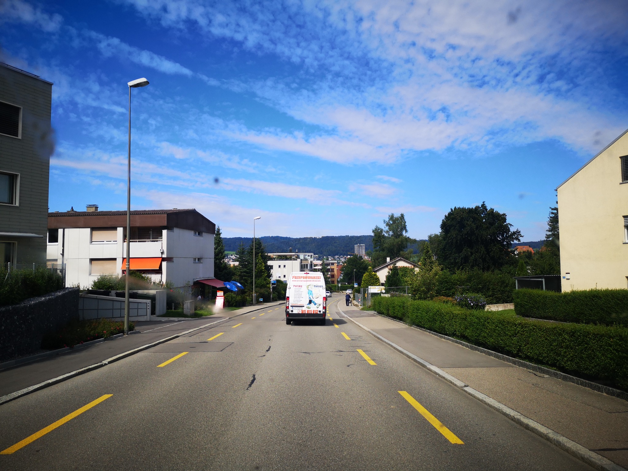 Biały bus firmy przeprowadzkowej z reklamą na karoserii, jadący ulicą w zabudowanym terenie w słoneczny dzień.