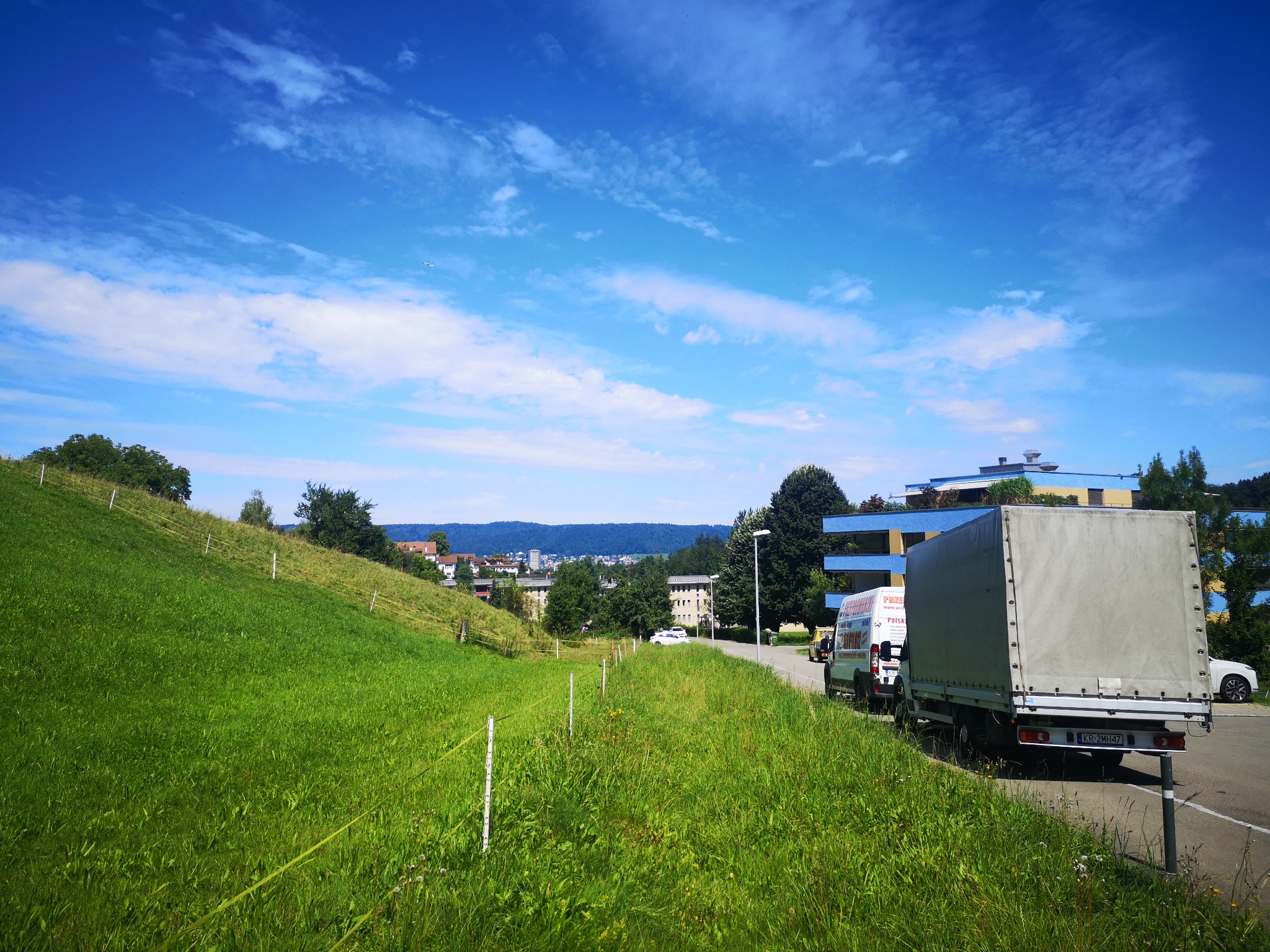 Dwa busy transportowe zaparkowane na poboczu drogi, z widokiem na zielone wzgórze i zabudowania miejskie w oddali pod błękitnym niebem.