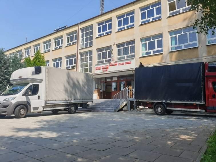 Dwa busy transportowe, biały i czerwony z czarną plandeką, zaparkowane przed budynkiem szkoły z szarymi schodami wejściowymi.