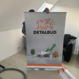 Rozwinięty roll-up reklamowy firmy budowlanej Detalbud z logo gryfa i hasłem 'Detale budują przewagę', prezentujący wizualizację domu i rosnący wykres słupkowy monet na pomarańczowym tle.