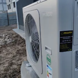Zewnętrzna jednostka klimatyzacyjna Hyundai na betonowej podstawie, widoczna naklejka firmy instalacyjnej i oznaczenie czynnika chłodniczego R32, w tle budynek w trakcie budowy.