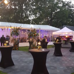 CZERSKA 8/10 SPÓŁKA Z OGRANICZONĄ ODPOWIEDZIALNOŚCIĄ - Plenerowa impreza cateringowa: wysokie stoliki z czarnymi pokrowcami, dekoracje kwiatowe z słonecznikami, oświetlony namiot w tle, goście wewnątrz.