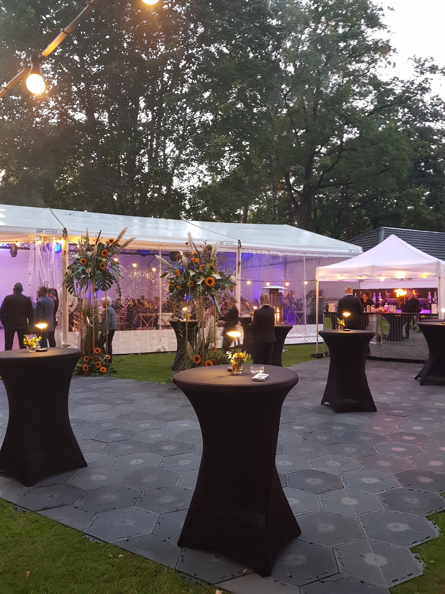 Plenerowa impreza cateringowa: wysokie stoliki z czarnymi pokrowcami, dekoracje kwiatowe z słonecznikami, oświetlony namiot w tle, goście wewnątrz.