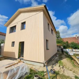 Dom w trakcie budowy z drewnianą elewacją, widoczne okna i drzwi, błękitne niebo z chmurami w tle. Na pierwszym planie materiały budowlane.