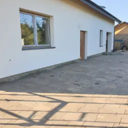 Nowoczesny dom jednorodzinny z jasną elewacją i nową kostką brukową na podwórku, widoczne okno z odbiciem drzew, rynna oraz budynek gospodarczy w tle.