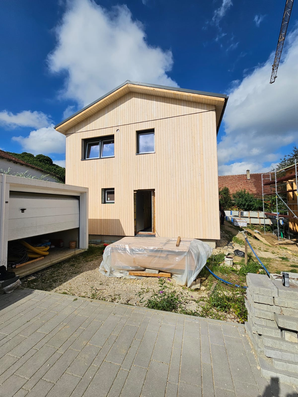 Dom w trakcie budowy z drewnianą elewacją, widoczne okna i otwarte drzwi. W tle błękitne niebo z chmurami i częściowo otwarty garaż.