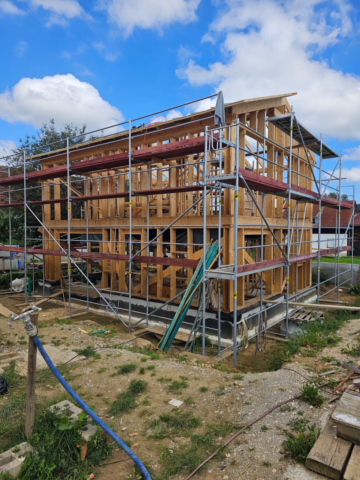Wznoszenie dwukondygnacyjnej drewnianej konstrukcji domu w systemie szkieletowym, osłoniętej rusztowaniami na tle błękitnego nieba z chmurami.