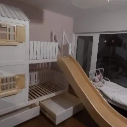 Białe łóżko piętrowe w kształcie domku z otwieranymi okiennicami i zjeżdżalnią, dodatkowo wysuwane szuflady na dole, obok regał z półkami.