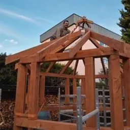 Konstrukcja drewnianej altany w trakcie budowy, z uśmiechniętym mężczyzną na dachu, widok na budynek w tle i stos drewna obok.