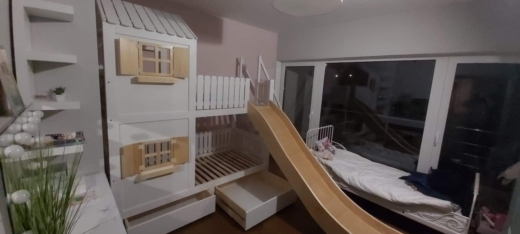 Białe łóżko piętrowe w kształcie domku z otwieranymi okiennicami i zjeżdżalnią, dodatkowo wysuwane szuflady na dole, obok regał z półkami.