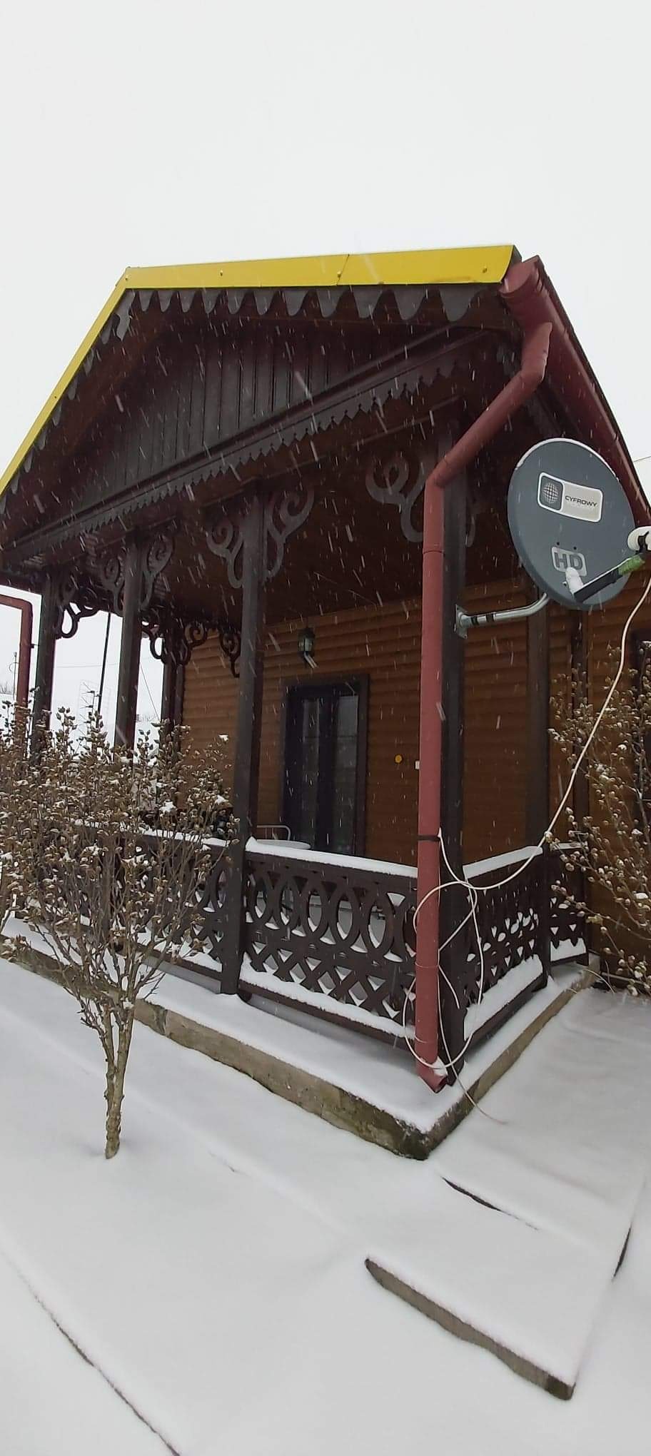 Drewniana altana z żółtym dachem i dekoracyjnymi elementami w zimowej scenerii, pokryta śniegiem, z widoczną anteną satelitarną.