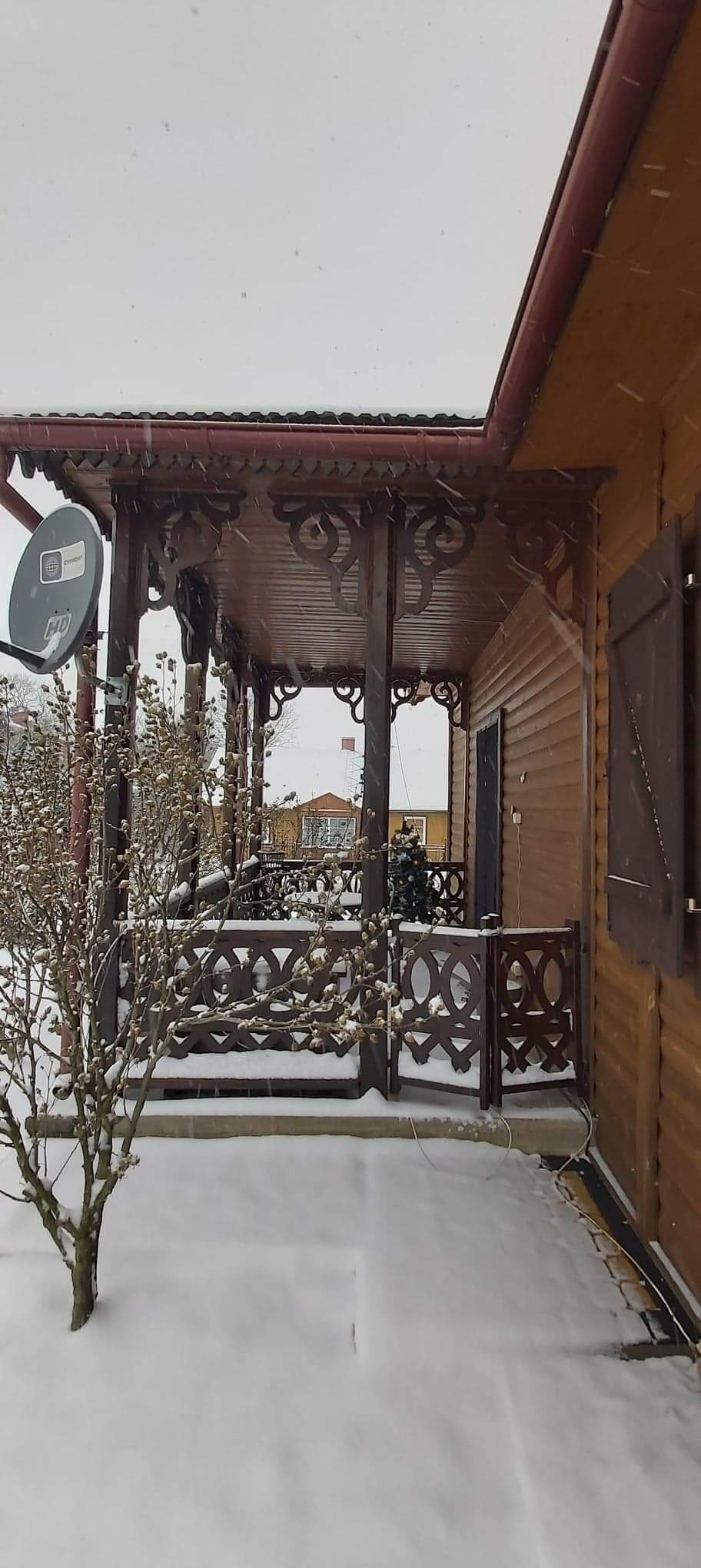 Zadaszony taras drewniany z dekoracyjnymi wycięciami, pokryty śniegiem podczas opadów, widoczna antena satelitarna i drzewo z pąkami.