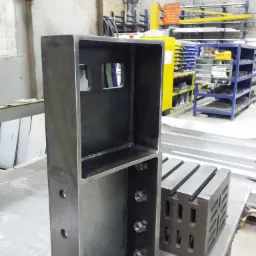 Surowa, metalowa konstrukcja spawana w kształcie prostopadłościanu z otworami i wewnętrzną półką, widoczne ślady spawania, obok kostka pomiarowa, tło warsztatowe z regałami i sprzętem.