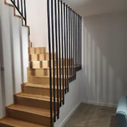 Drewniane schody z nowoczesną balustradą z pionowych, czarnych prętów, oświetlone punktowym światłem, z fragmentem jasnej podłogi i sofy w tle.