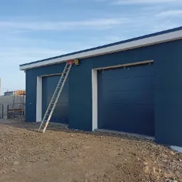 Nowoczesny budynek garażowy z dwoma bramami w kolorze grafitowym, otynkowany na ciemno, z białą podbitką dachową i drabiną opartą o elewację, na tle błękitnego nieba.