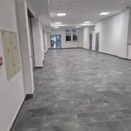 Wyremontowany korytarz w budynku Zgorzelec Francuska po rewitalizacji, z szarymi płytkami podłogowymi imitującymi kamień, białymi ścianami, sufitem podwieszanym z oświetleniem LED i drzwiami...