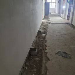 Przygotowanie do instalacji ogrzewania podłogowego: wykopany rów wzdłuż ściany, widoczne bloczki betonowe, surowy beton na podłodze i fragmenty instalacji elektrycznej pod sufitem.