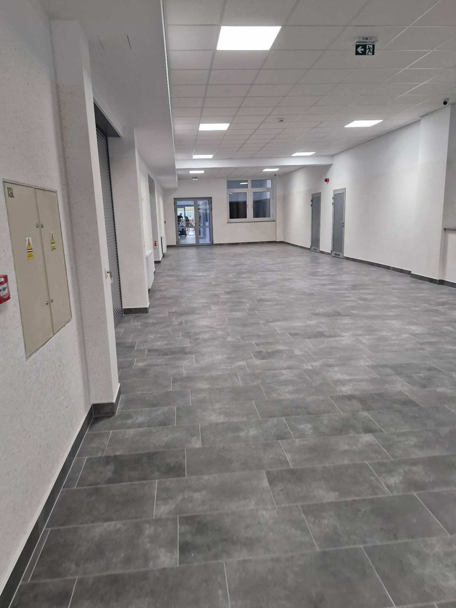 Wyremontowany korytarz w budynku Zgorzelec Francuska po rewitalizacji, z szarymi płytkami podłogowymi imitującymi kamień, białymi ścianami, sufitem podwieszanym z oświetleniem LED i drzwiami...