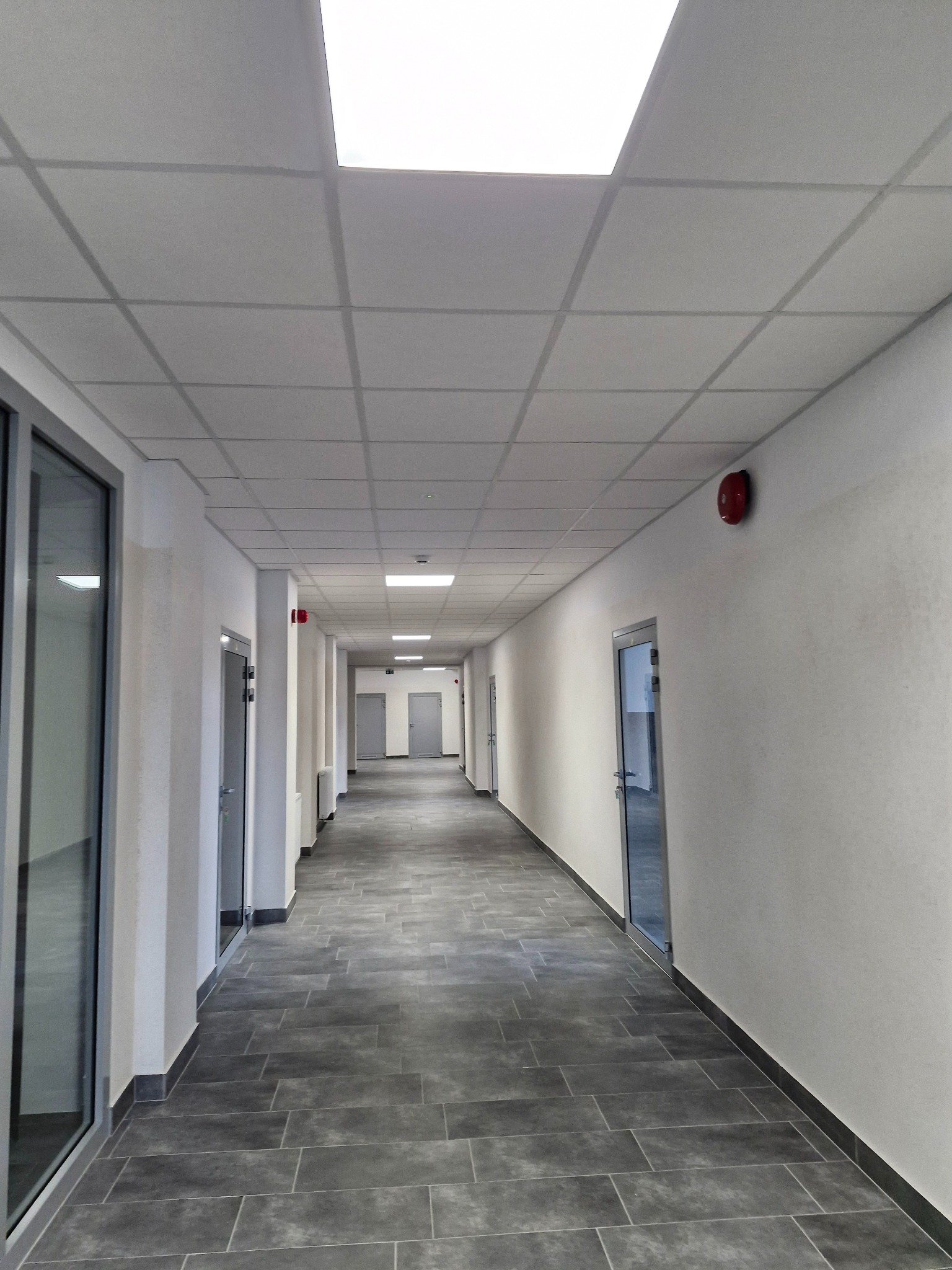 Długi korytarz biurowy z szarymi płytkami podłogowymi ułożonymi w cegiełkę, białymi ścianami, sufitem podwieszanym z kwadratowymi panelami i oświetleniem oraz czerwonym alarmem przeciwpożarowym...
