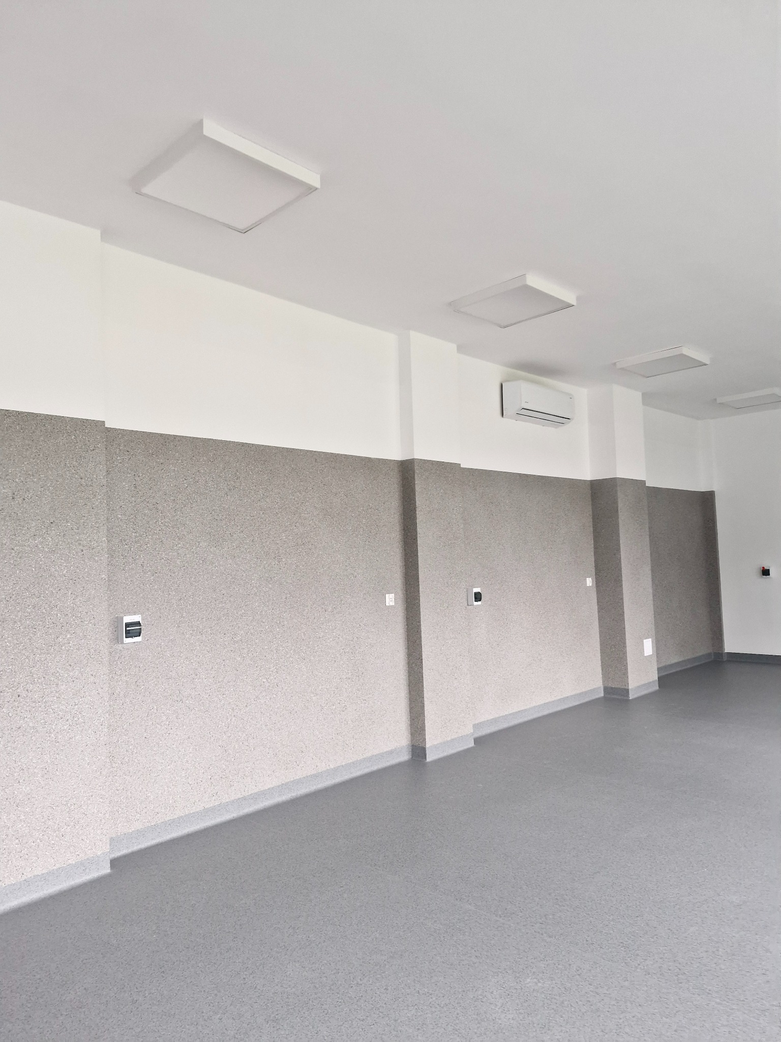 Wnętrze pomieszczenia z szarą podłogą i ścianami wykończonymi panelami do połowy wysokości, widoczne kwadratowe lampy sufitowe, klimatyzator oraz gniazdka elektryczne.