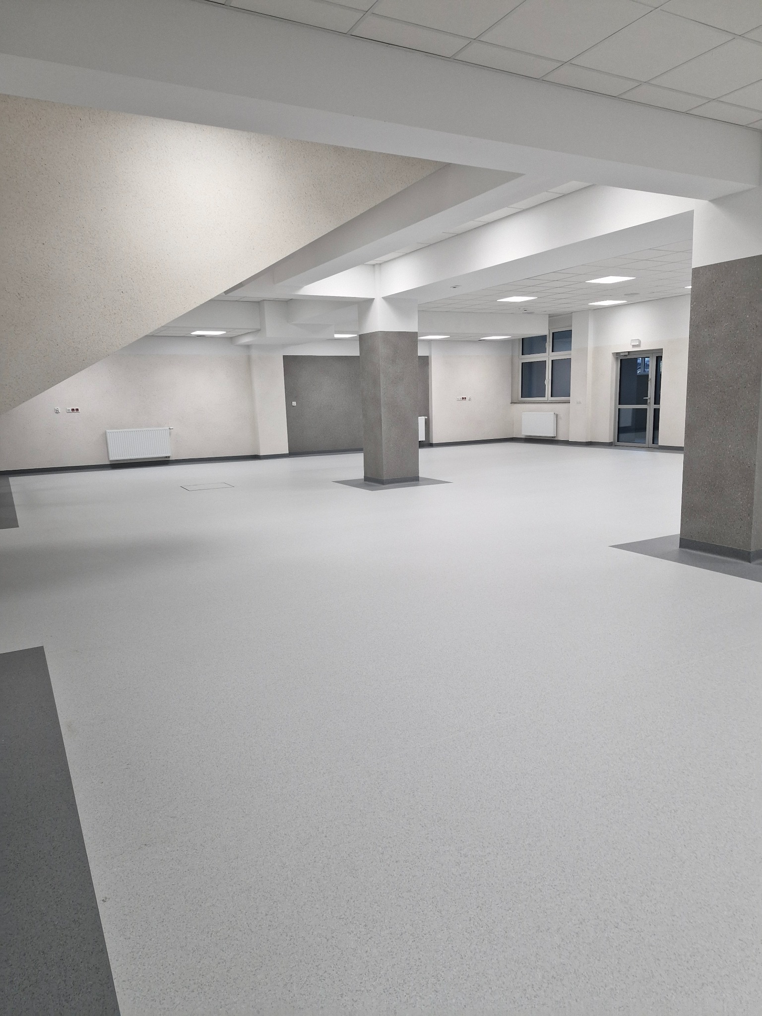 Przestronne, jasne wnętrze z szarą podłogą, grzejnikiem na ścianie i kolumnami w kolorze szarym, widoczne okna i drzwi.
