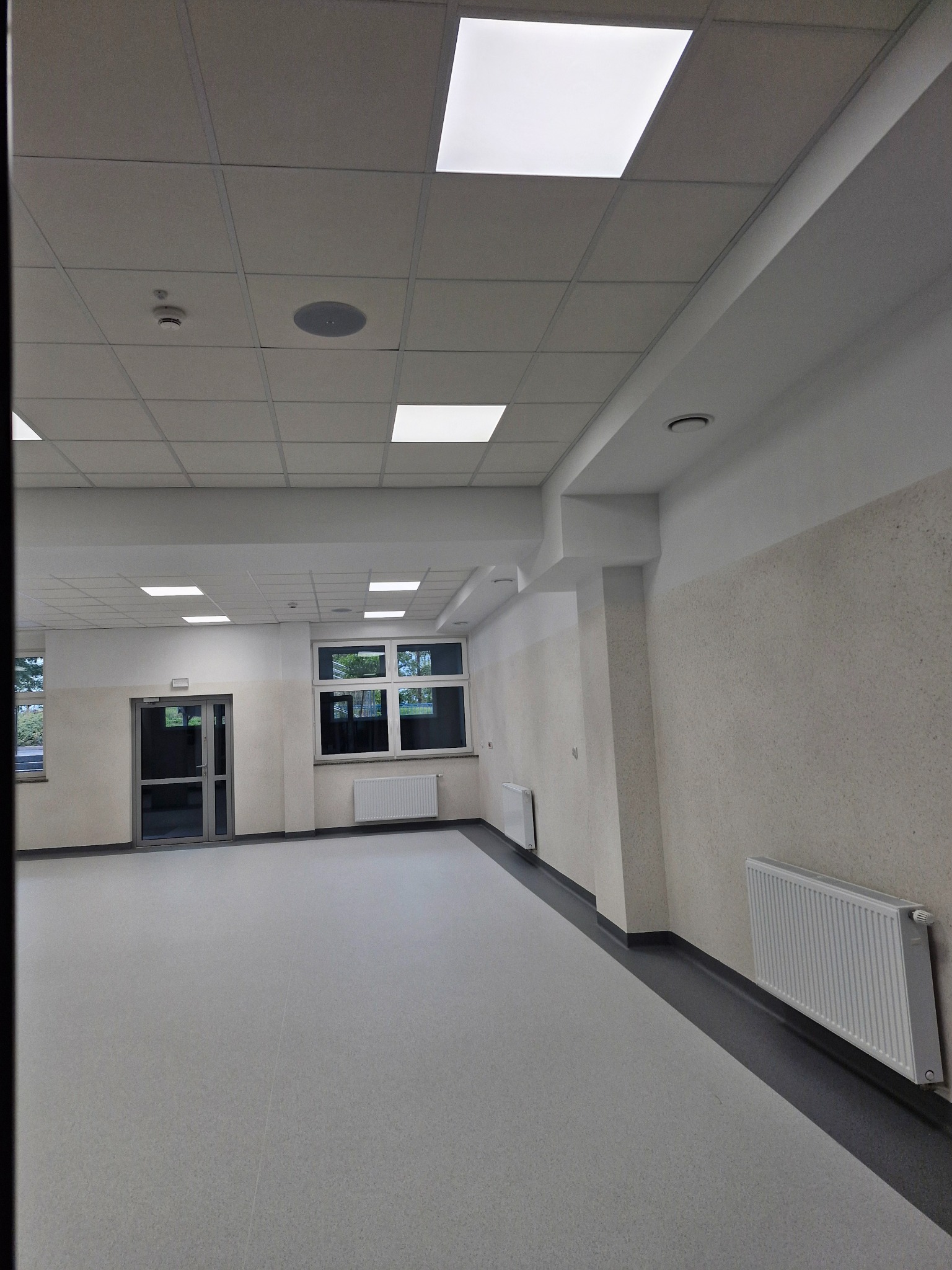 Wnętrze wykończonego pomieszczenia z szarymi panelami podłogowymi, białymi grzejnikami, oknami i sufitem kasetonowym z oświetleniem LED.
