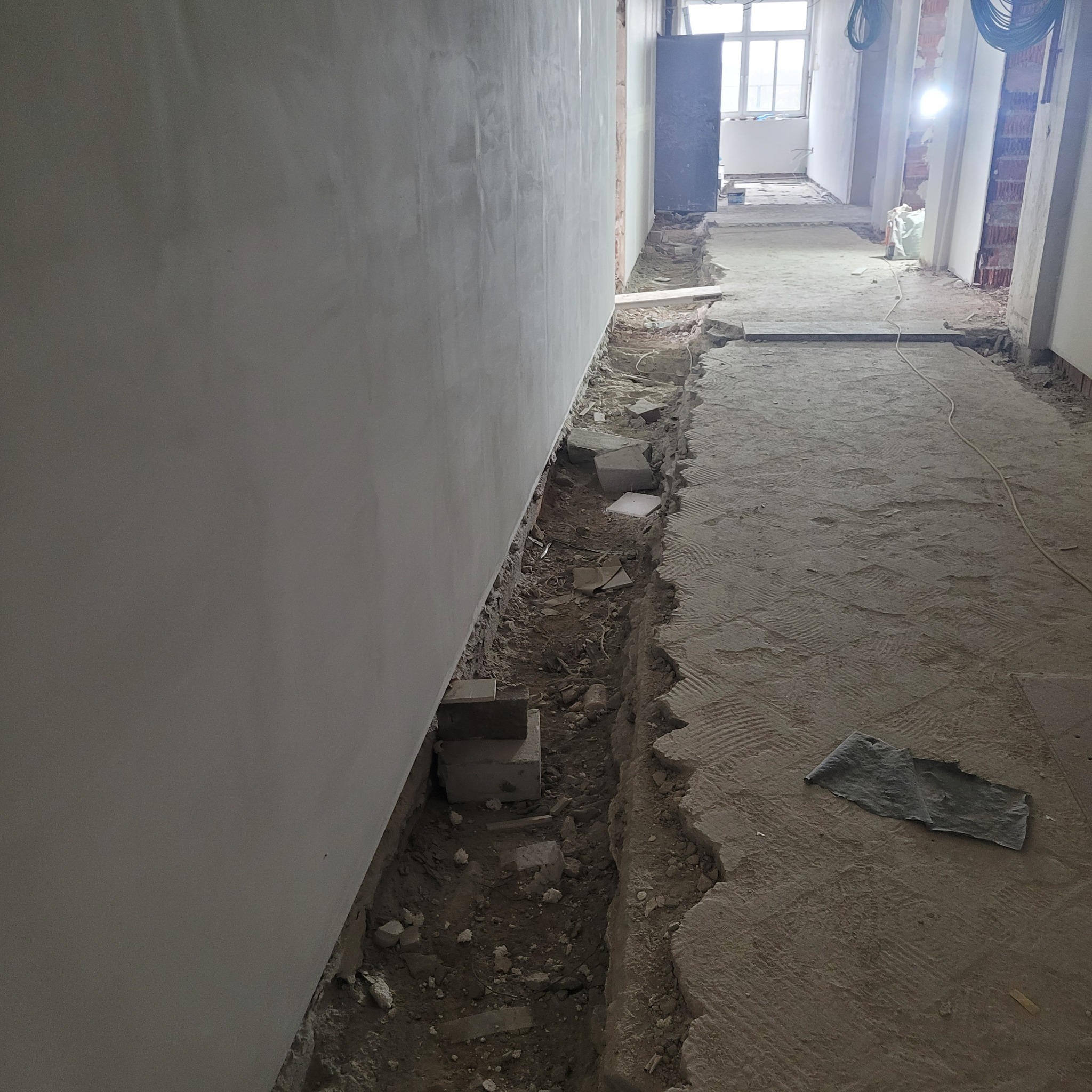 Przygotowanie do instalacji ogrzewania podłogowego: wykopany rów wzdłuż ściany, widoczne bloczki betonowe, surowy beton na podłodze i fragmenty instalacji elektrycznej pod sufitem.