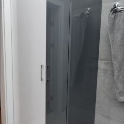 Krzysztof Rozum - Nowoczesna szafa wnękowa z białymi drzwiami i szarą, połyskującą powierzchnią, z widocznym fragmentem łazienki w tle.