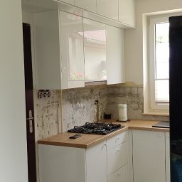 Krzysztof Rozum - Wnętrze kuchni w trakcie remontu: białe, lakierowane szafki wiszące i dolne z drewnianym blatem, widoczne ślady po starych płytkach na ścianie, płyta gazowa na blacie.