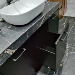 Krzysztof Rozum - Łazienka z umywalką nablatową i czarną szafką z otwartą szufladą na tle marmurowej ściany i podłogi.