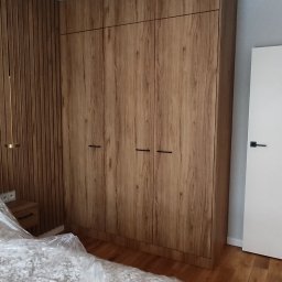 Krzysztof Rozum - Wysoka szafa z frontami imitującymi drewno, z czarnymi uchwytami, dopasowana do ściany z drewnianymi panelami w sypialni, łóżko przykryte folią ochronną.