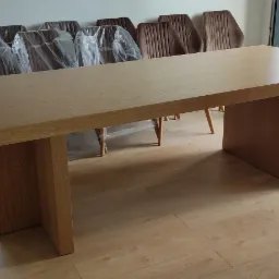 Jasny, prostokątny st&oacute;ł z litego drewna z grubym blatem i nogami, ustawiony w nowoczesnym wnętrzu na podłodze z paneli, z krzesłami obitymi brązowym welurem, niekt&oacute;re jeszcze w folii ochronnej...