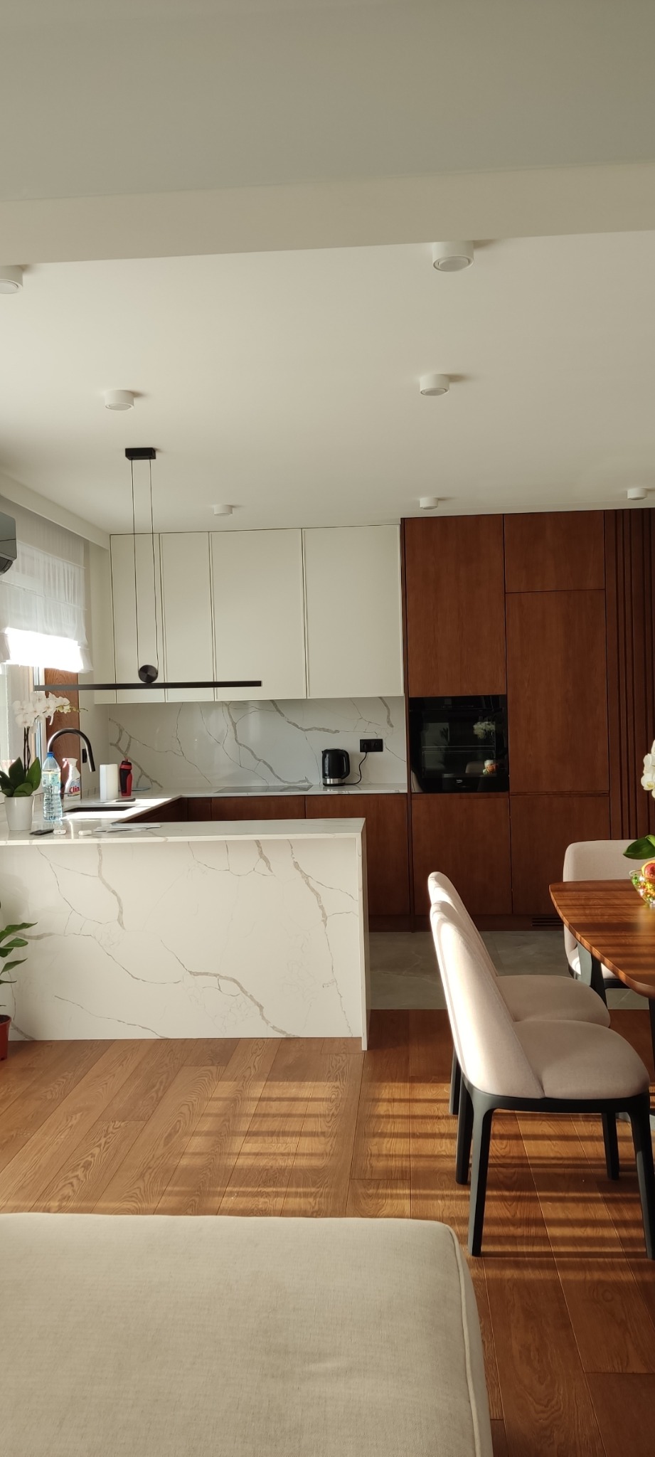 Nowoczesna kuchnia z białymi i drewnianymi szafkami, blatem z białego marmuru i drewnianą podłogą, oświetlona światłem słonecznym.