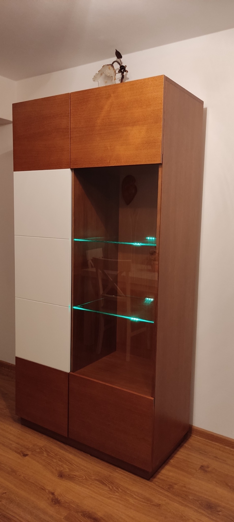 Dwukolorowa witryna z drewnianymi i białymi frontami, szklane półki podświetlone ledami, na górze dekoracyjny bibelot.