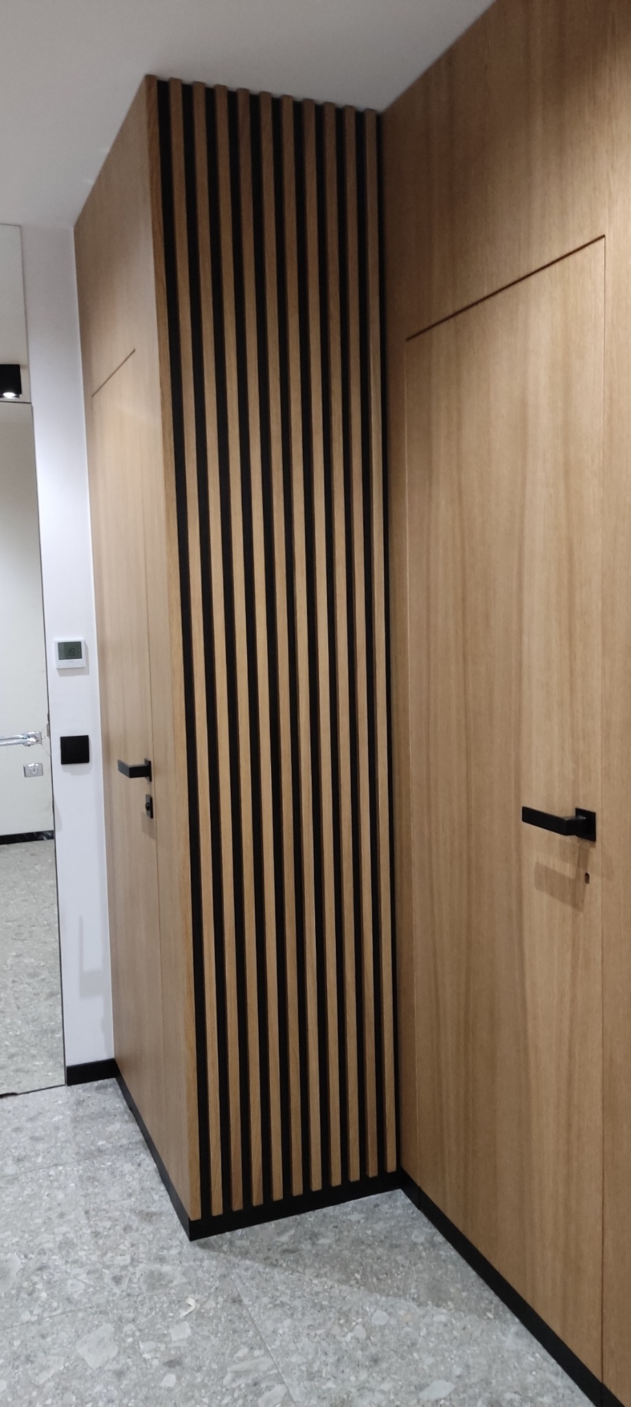 Drewniana zabudowa w przedpokoju z lamelami i drzwiami, czarne klamki, szara podłoga, widok na lustro i białe ściany w tle.