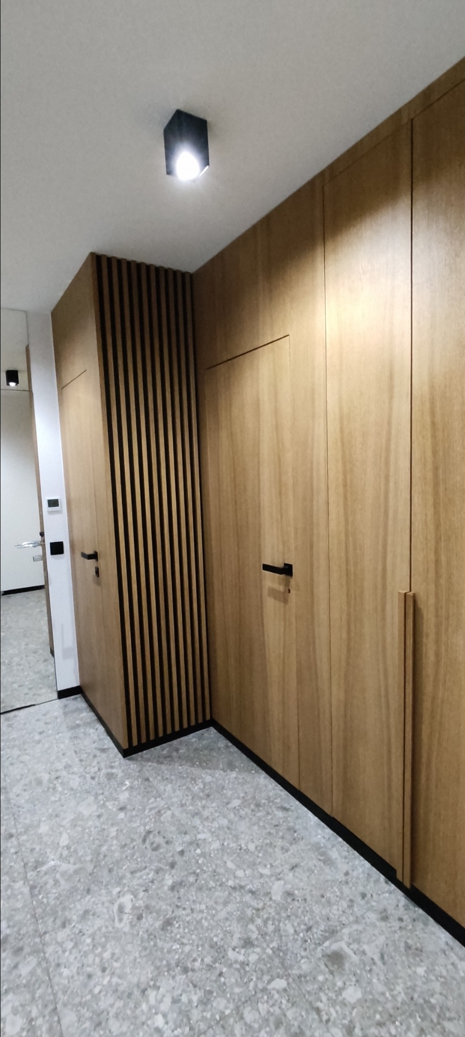 Wnętrze przedpokoju z zabudową szafy z drewnianymi frontami i pionowymi czarnymi listwami dekoracyjnymi, czarne uchwyty, szara posadzka z wzorem lastryko.