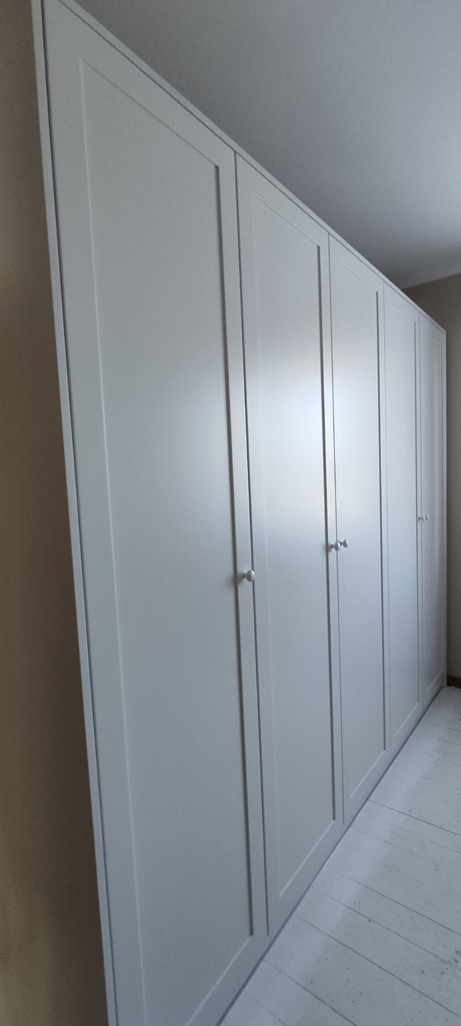 Biała szafa wnękowa z czterema drzwiami, z okrągłymi uchwytami, zamontowana od podłogi do sufitu w jasnym pomieszczeniu.