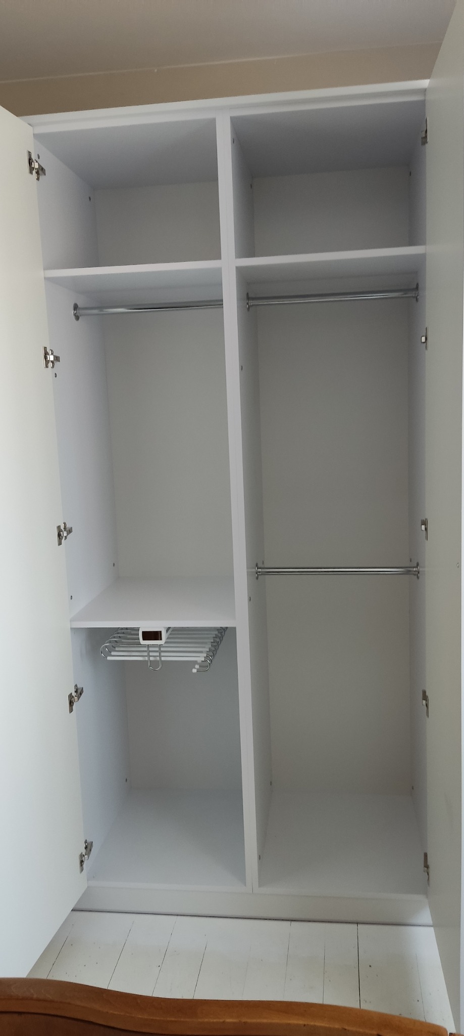 Wnętrze białej, dwudrzwiowej szafy z półkami i drążkami na ubrania, widoczne zawiasy i wysuwany wieszak na spodnie.
