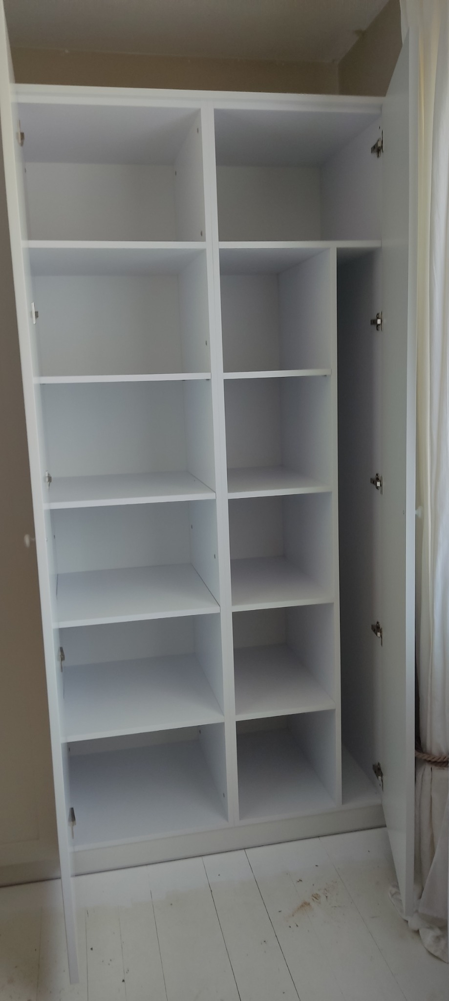 Biała, pusta szafa z otwartymi drzwiami, widoczne półki o różnej szerokości, minimalistyczny design.