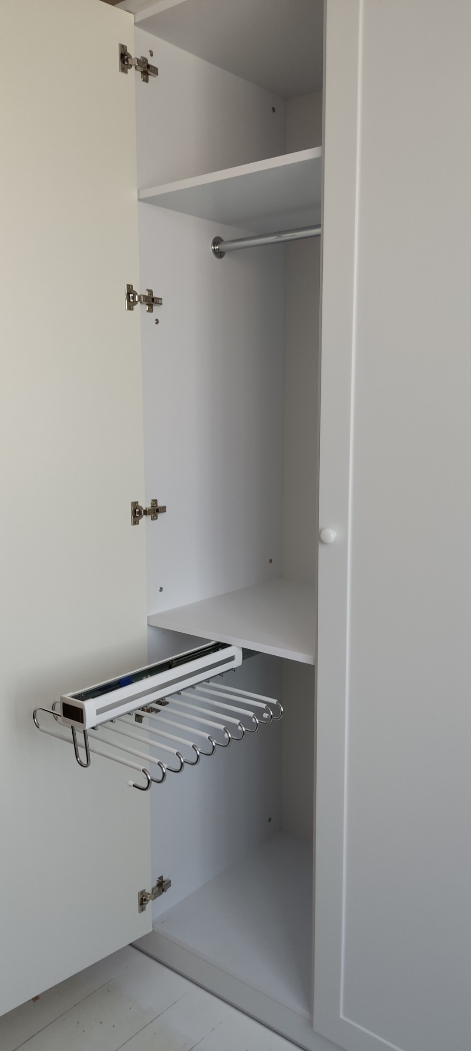 Wnętrze białej szafy z zamontowanym wysuwanym wieszakiem na spodnie i półkami.