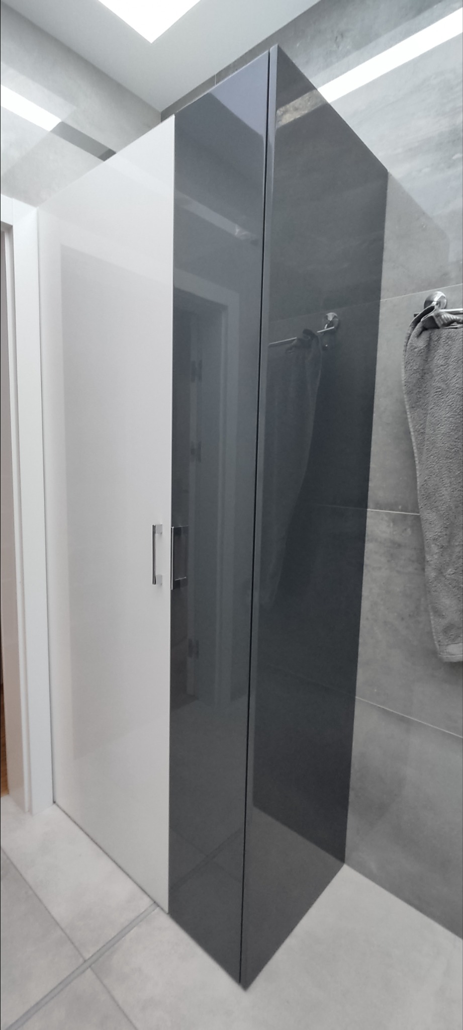 Nowoczesna szafa wnękowa z białymi drzwiami i szarą, połyskującą powierzchnią, z widocznym fragmentem łazienki w tle.