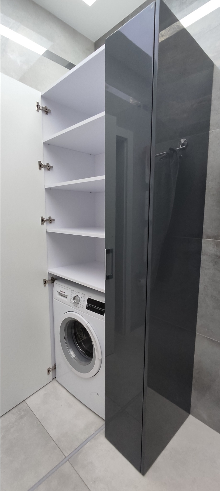 Biało-szara szafa na wymiar z otwartymi drzwiami, kryjąca pralkę Bosch w nowoczesnej łazience z szarymi płytkami.