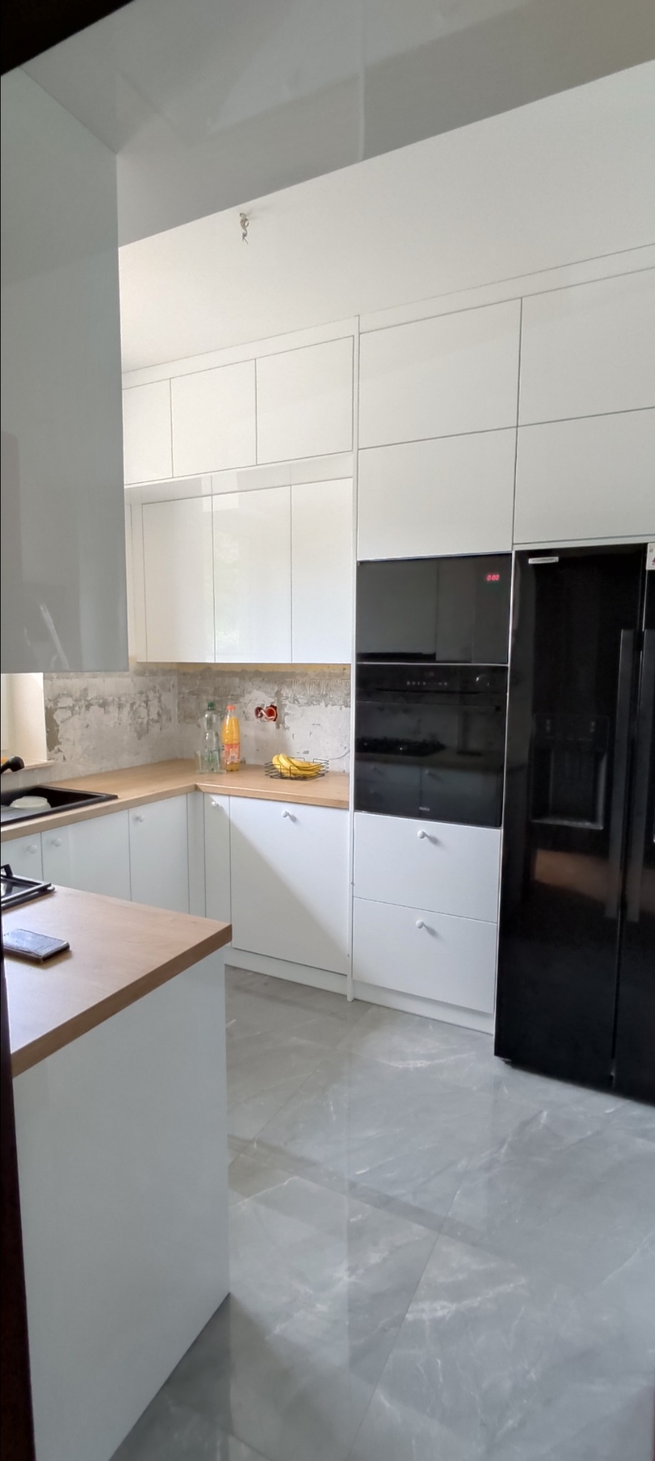 Jasna kuchnia z białymi szafkami i drewnianym blatem, widoczna podczas remontu, ściana częściowo bez wykończenia, na blacie owoce i napoje.